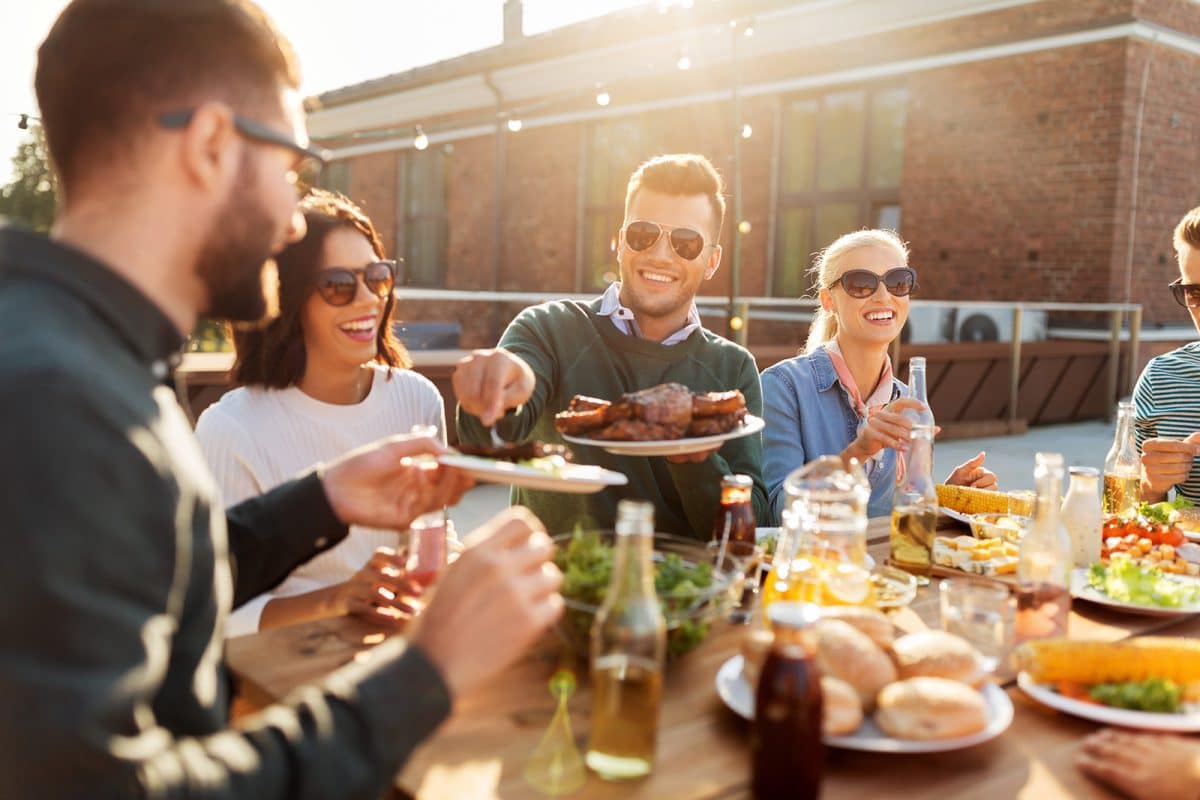 A group of people eating a meal together, with many dishes in the middle and a sun setting behind a brick building behind the group