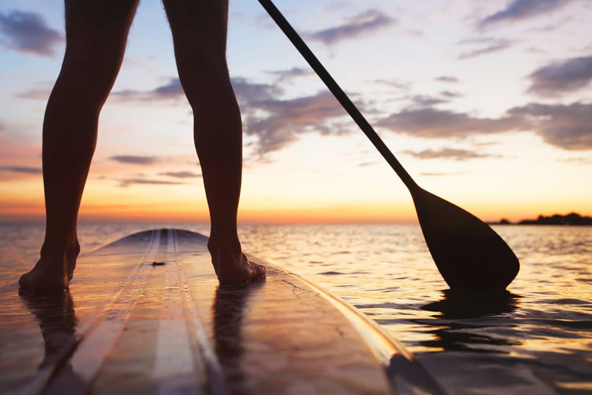 A paddle boarder at sunset, pictured from the knees down, showing their paddle in the water